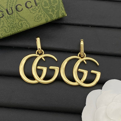 Gold pendants with double G design on a black surface, Gucci box in the background.